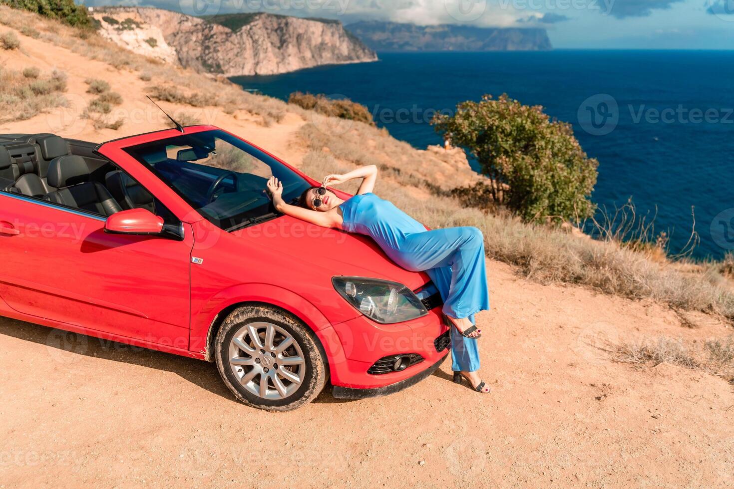 A woman is laying on top of a red convertible car. The car is parked on a dirt road near the ocean. The woman is wearing a blue dress and she is enjoying the moment. photo