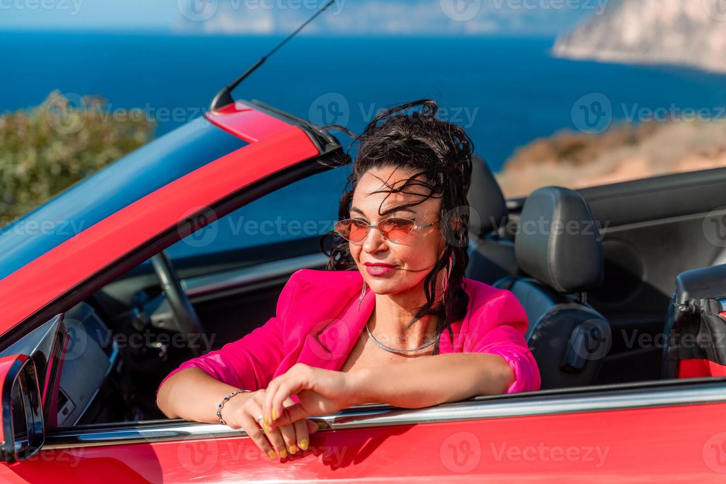 A woman in a pink jacket is sitting in a red convertible. She is wearing sunglasses and has her hand on her hip. Scene is relaxed and carefree. photo