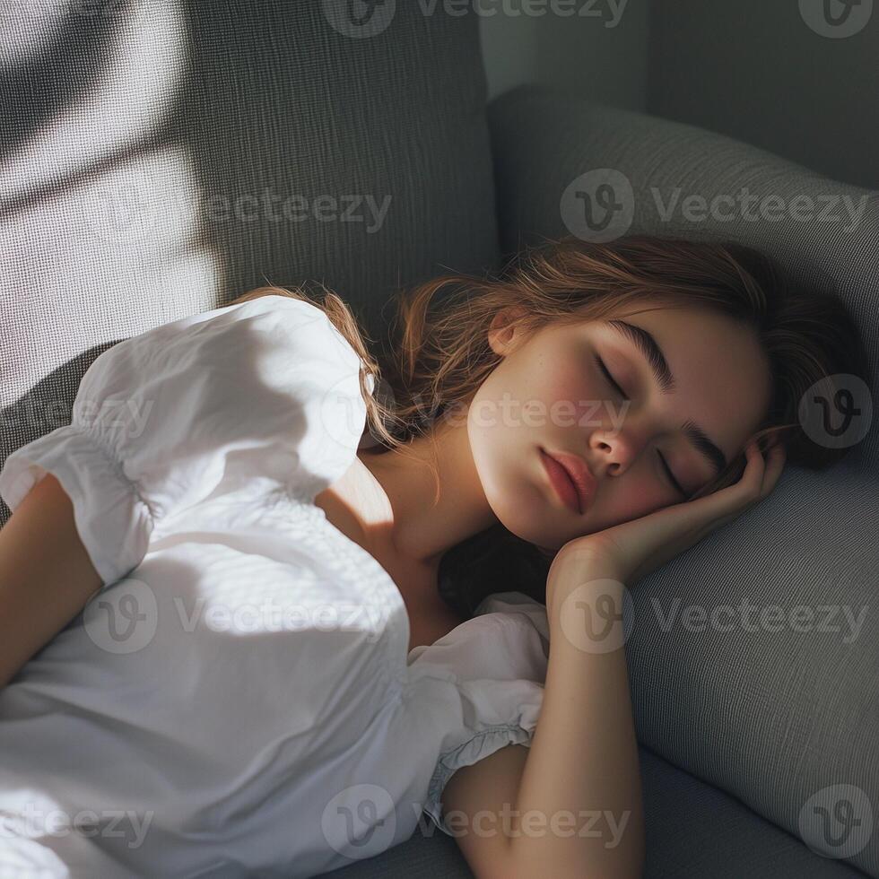 A young woman in a white outfit sleeps on a grey sofa, with soft light casting shadows, creating a calm atmosphere. photo