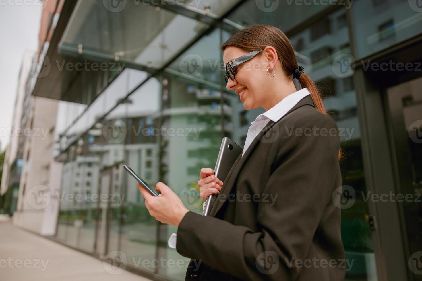 A confident businesswoman holds a tablet and documents, smiling outside a modern office building photo