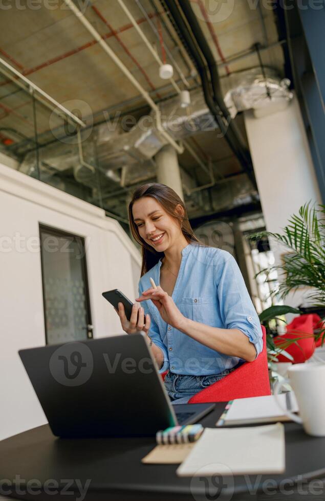 A young woman is happily working remotely from a modern office space, using a laptop and phone photo