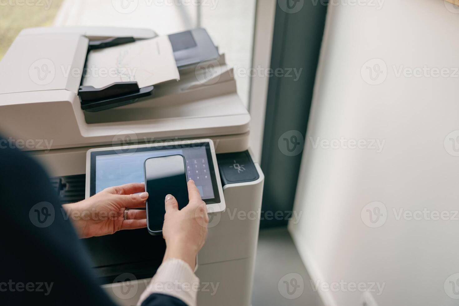 A person uses a smartphone to operate a modern printer in a corporate office setting photo
