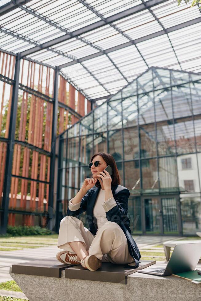 A businesswoman is working outdoors in an urban setting, talking on the phone and using a laptop photo