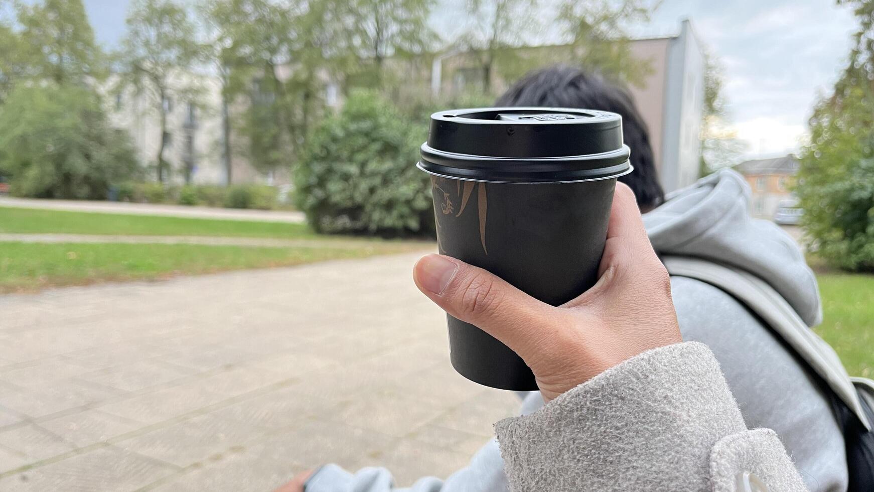 A hand holding a black coffee cup outdoors with a unrecognized man in background in Autumn season photo