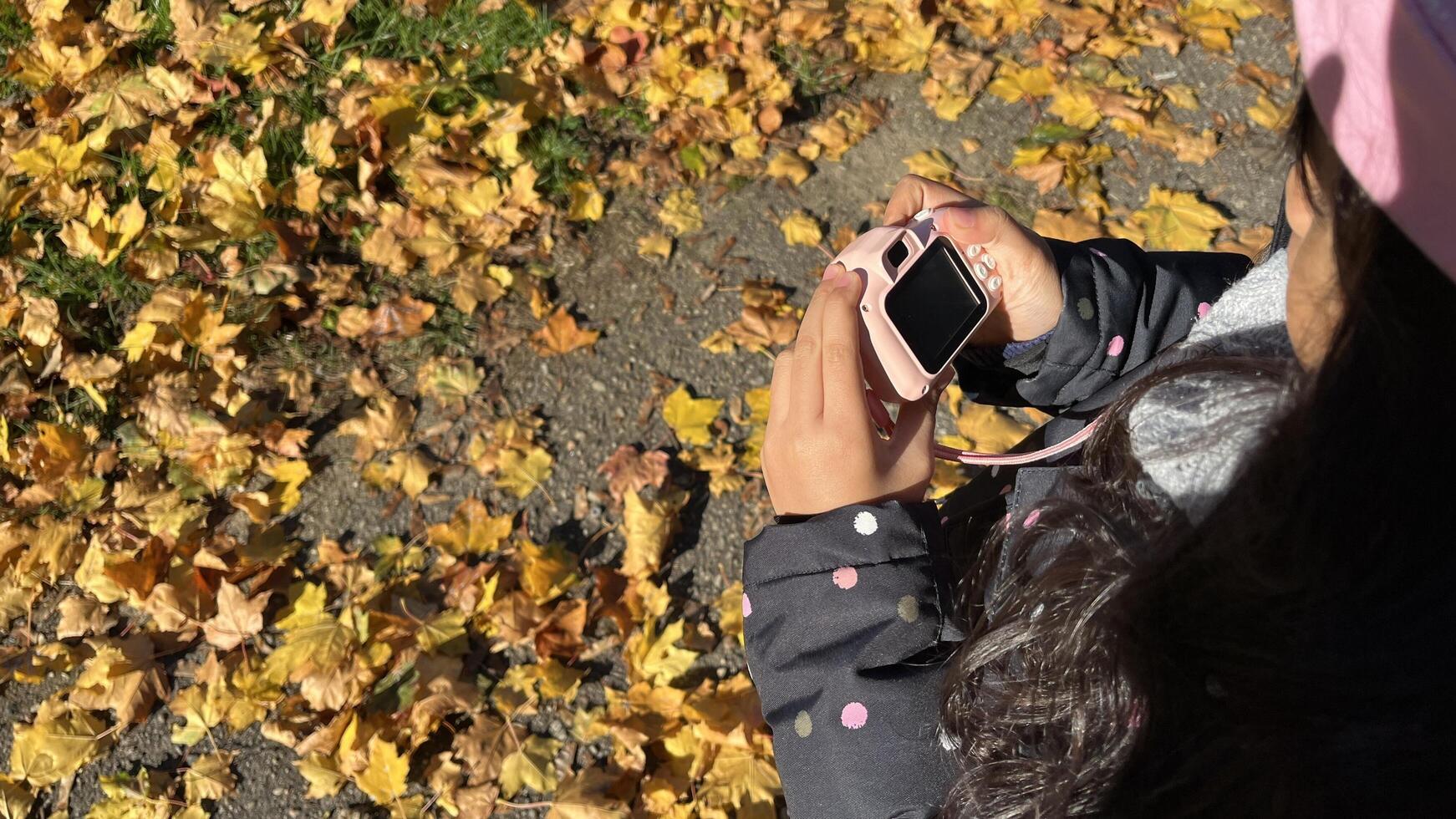 Beautiful Indonesian little girl holding mini kid camera in autumn season wearing padded jacket and pink beanie. Figure viewed from behind photo