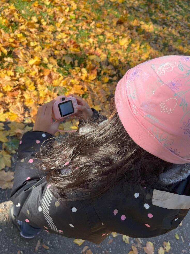 Beautiful Indonesian little girl holding mini kid camera in autumn season wearing padded jacket and pink beanie. Figure viewed from behind photo
