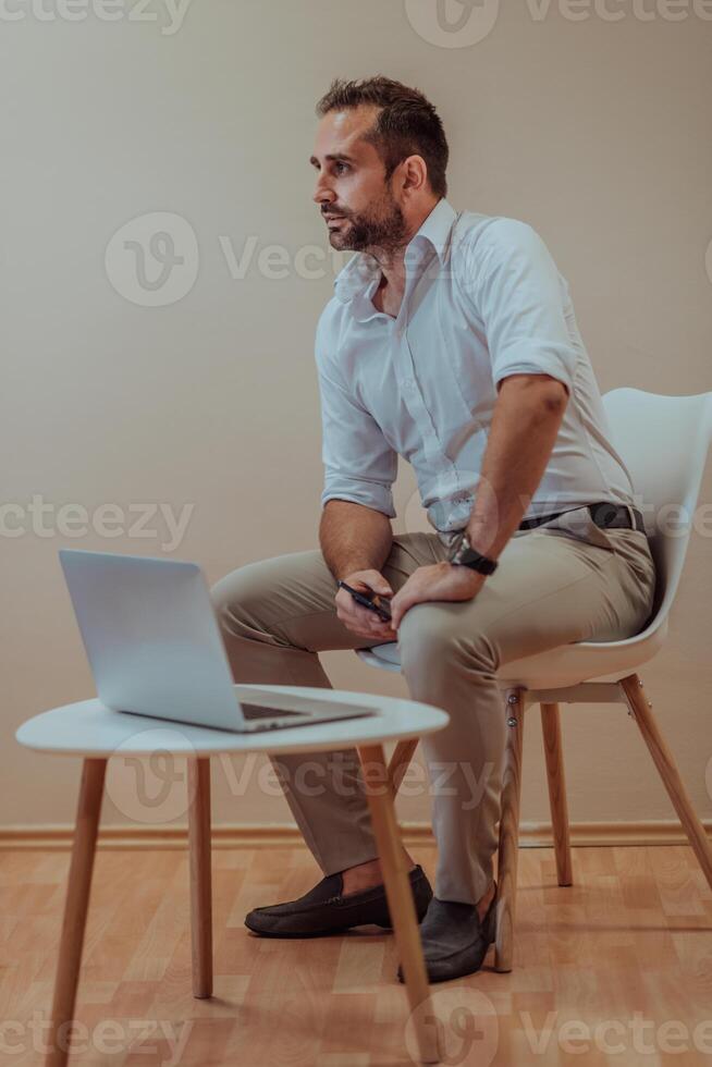 A confident businessman sitting and using laptop with a determined expression, while a beige background enhances the professional atmosphere, showcasing his productivity and expertise. photo