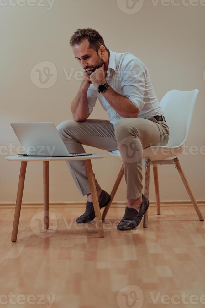 A confident businessman sitting and using laptop with a determined expression, while a beige background enhances the professional atmosphere, showcasing his productivity and expertise. photo