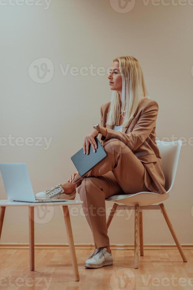 A professional businesswoman sits on a chair, surrounded by a serene beige background, diligently working on her laptop, showcasing dedication and focus in her pursuit of success photo