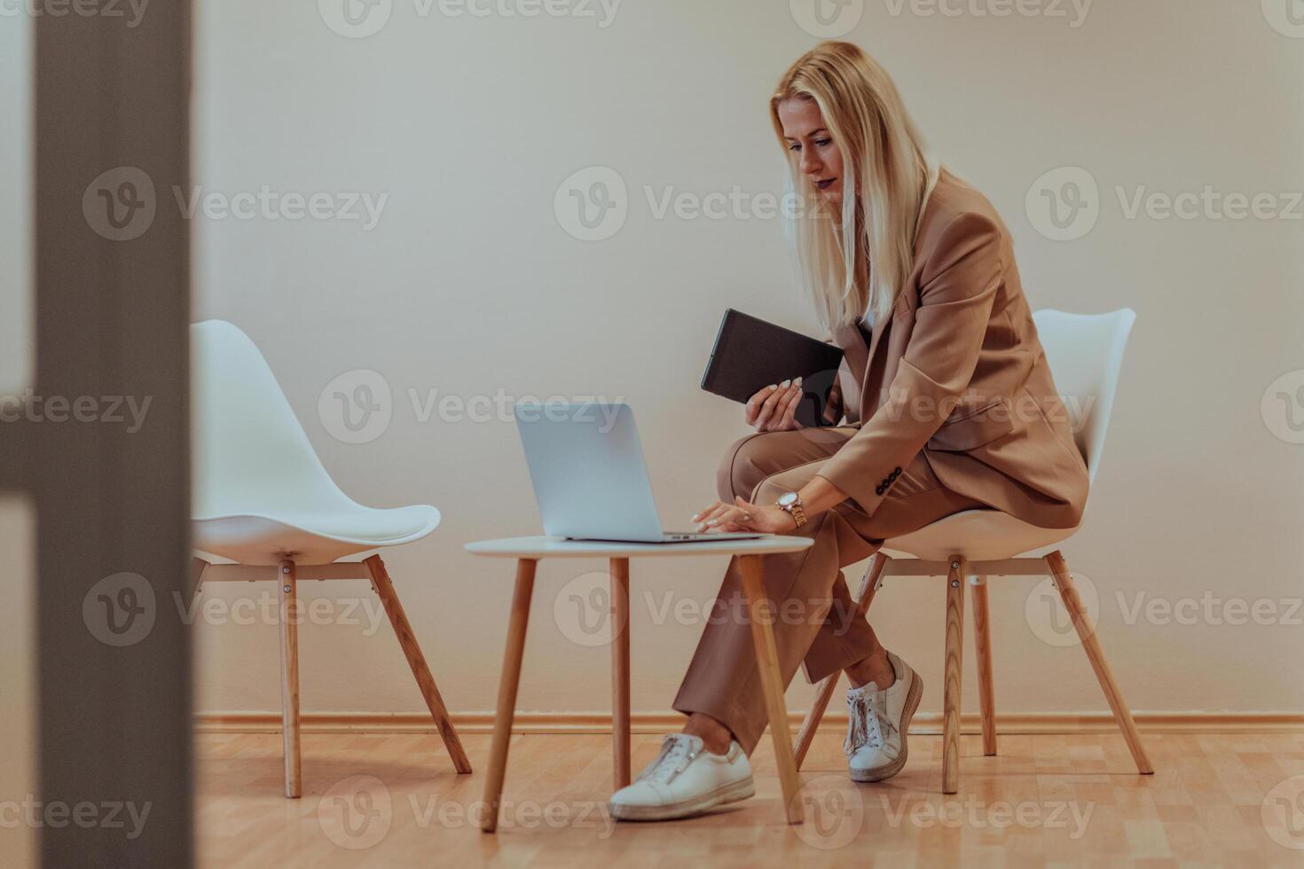 A professional businesswoman sits on a chair, surrounded by a serene beige background, diligently working on her laptop, showcasing dedication and focus in her pursuit of success photo