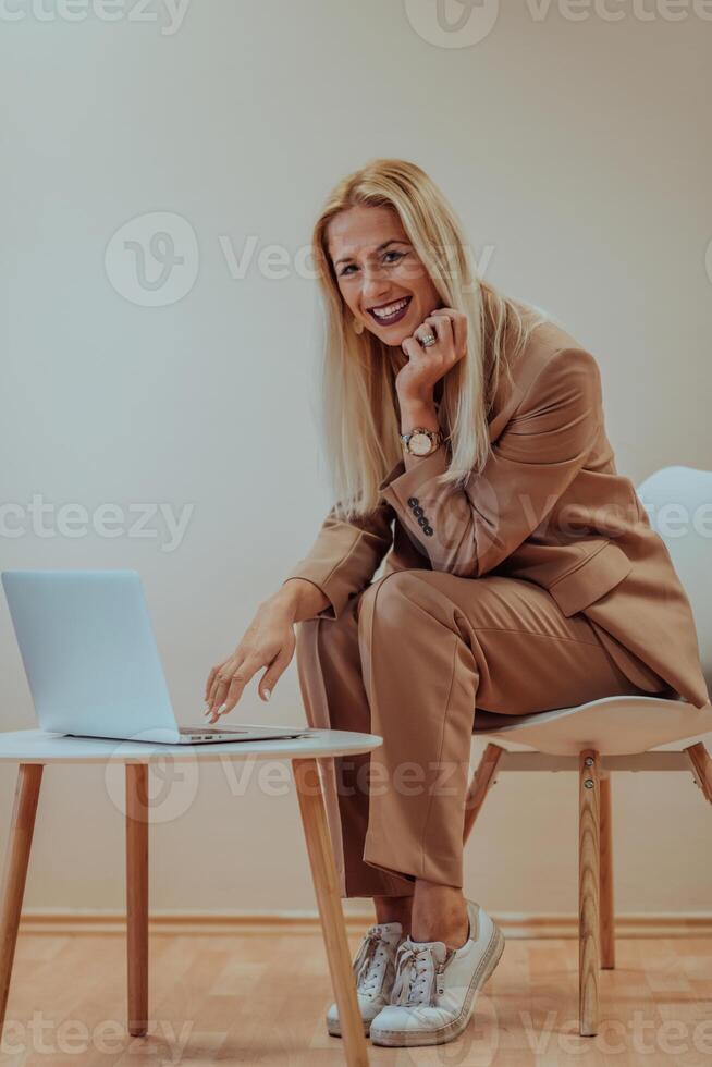 A professional businesswoman sits on a chair, surrounded by a serene beige background, diligently working on her laptop, showcasing dedication and focus in her pursuit of success photo