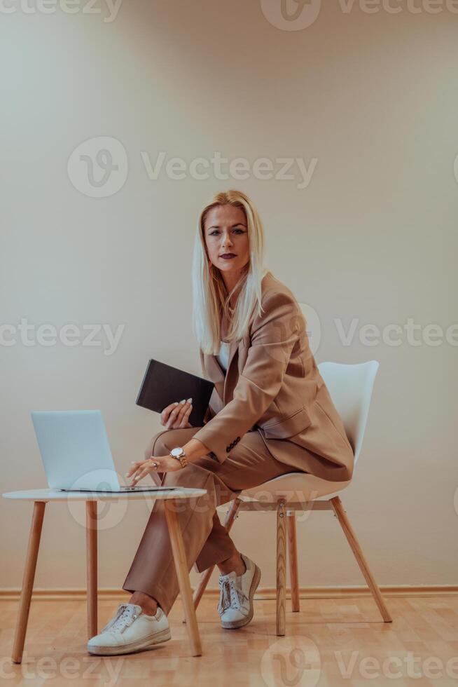 A professional businesswoman sits on a chair, surrounded by a serene beige background, diligently working on her laptop, showcasing dedication and focus in her pursuit of success photo