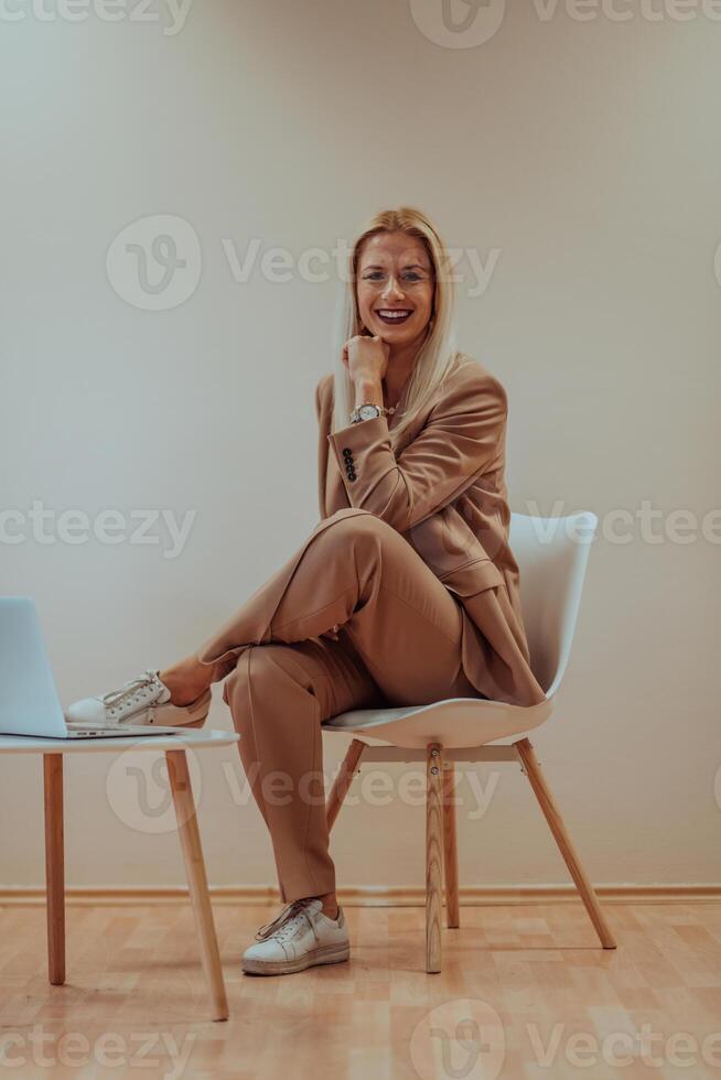 A professional businesswoman sits on a chair, surrounded by a serene beige background, diligently working on her laptop, showcasing dedication and focus in her pursuit of success photo