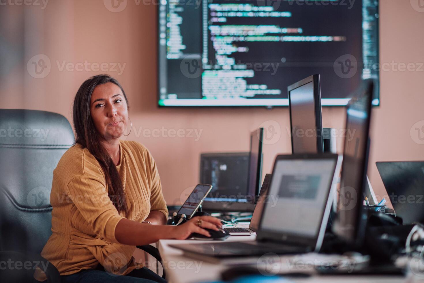 A businesswoman sitting in a programmer's office surrounded by computers, showing her expertise and dedication to technology. photo