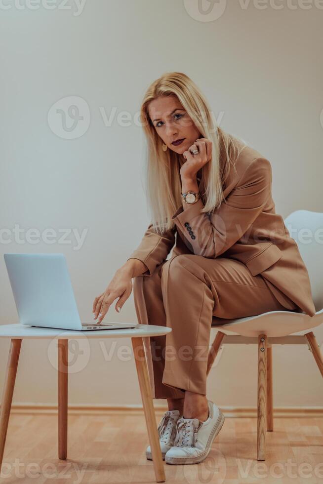 A professional businesswoman sits on a chair, surrounded by a serene beige background, diligently working on her laptop, showcasing dedication and focus in her pursuit of success photo