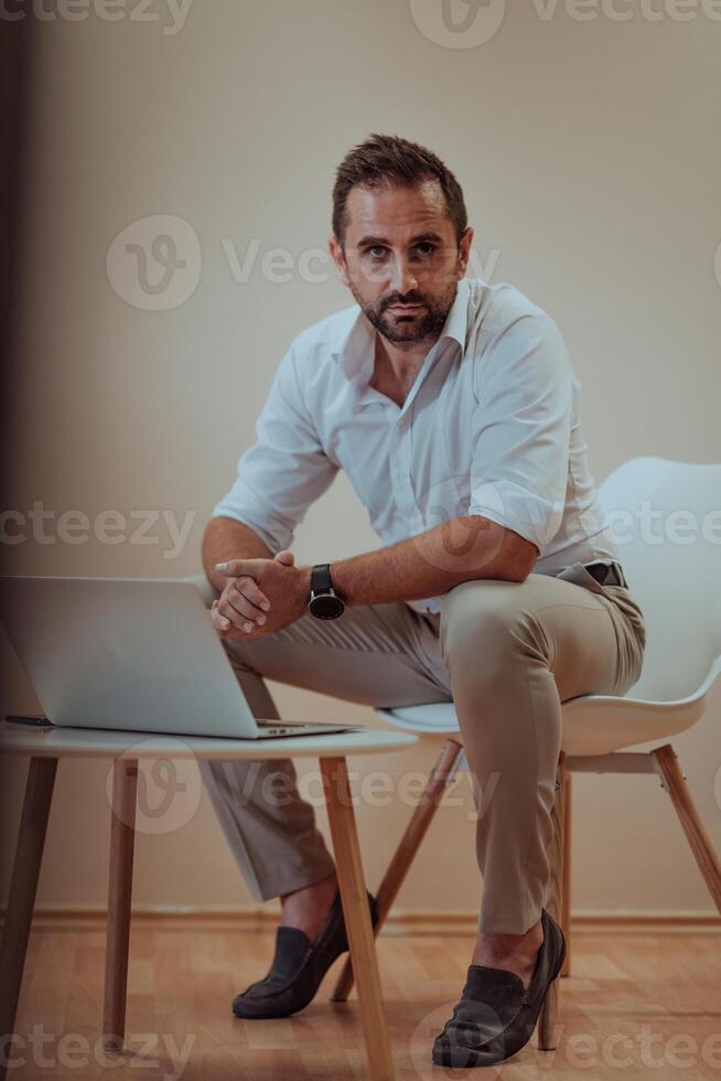 A confident businessman sitting and using laptop with a determined expression, while a beige background enhances the professional atmosphere, showcasing his productivity and expertise. photo