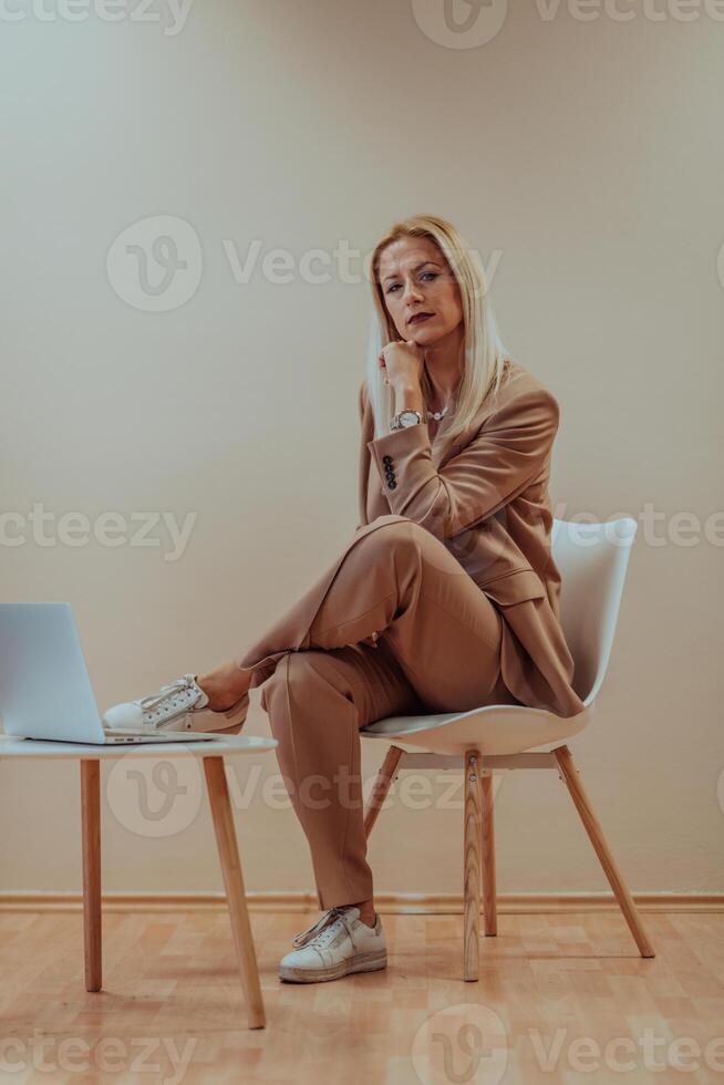 A professional businesswoman sits on a chair, surrounded by a serene beige background, diligently working on her laptop, showcasing dedication and focus in her pursuit of success photo