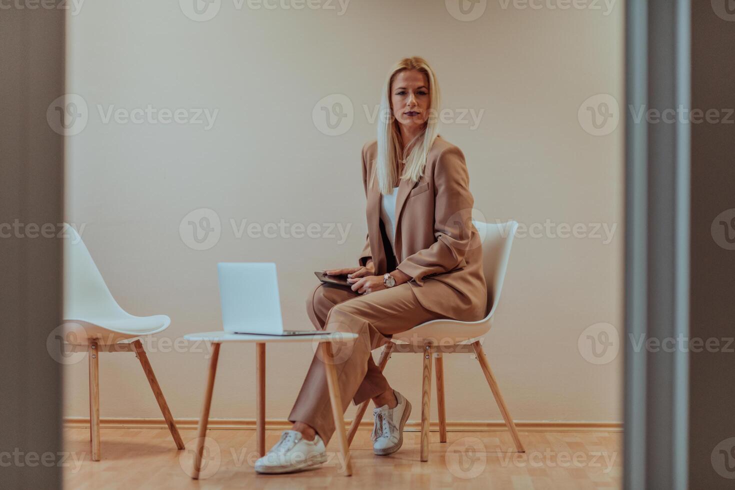 A professional businesswoman sits on a chair, surrounded by a serene beige background, diligently working on her laptop, showcasing dedication and focus in her pursuit of success photo