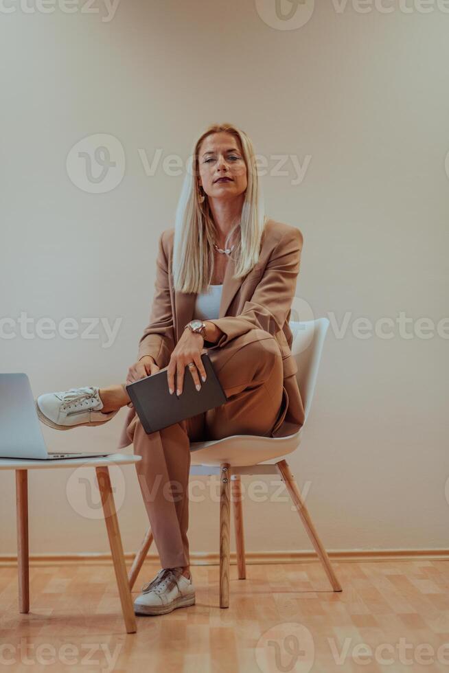A professional businesswoman sits on a chair, surrounded by a serene beige background, diligently working on her laptop, showcasing dedication and focus in her pursuit of success photo