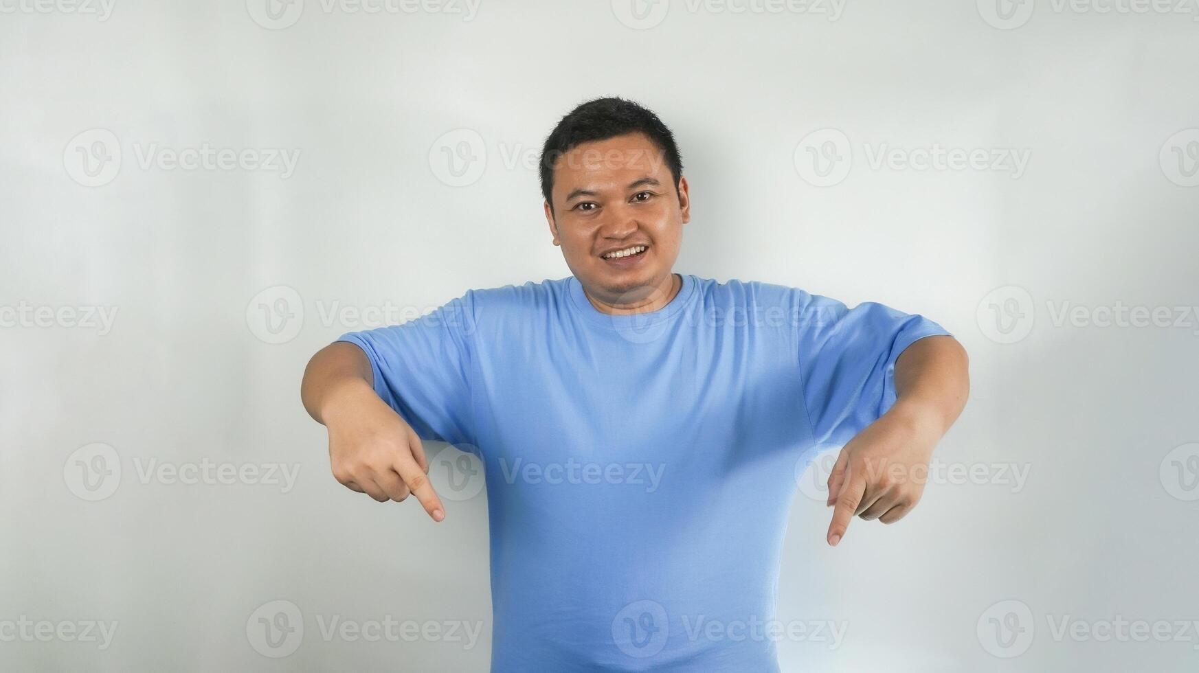 A young Indonesian man wearing a blue t-shirt with a happy face pointing downwards photo