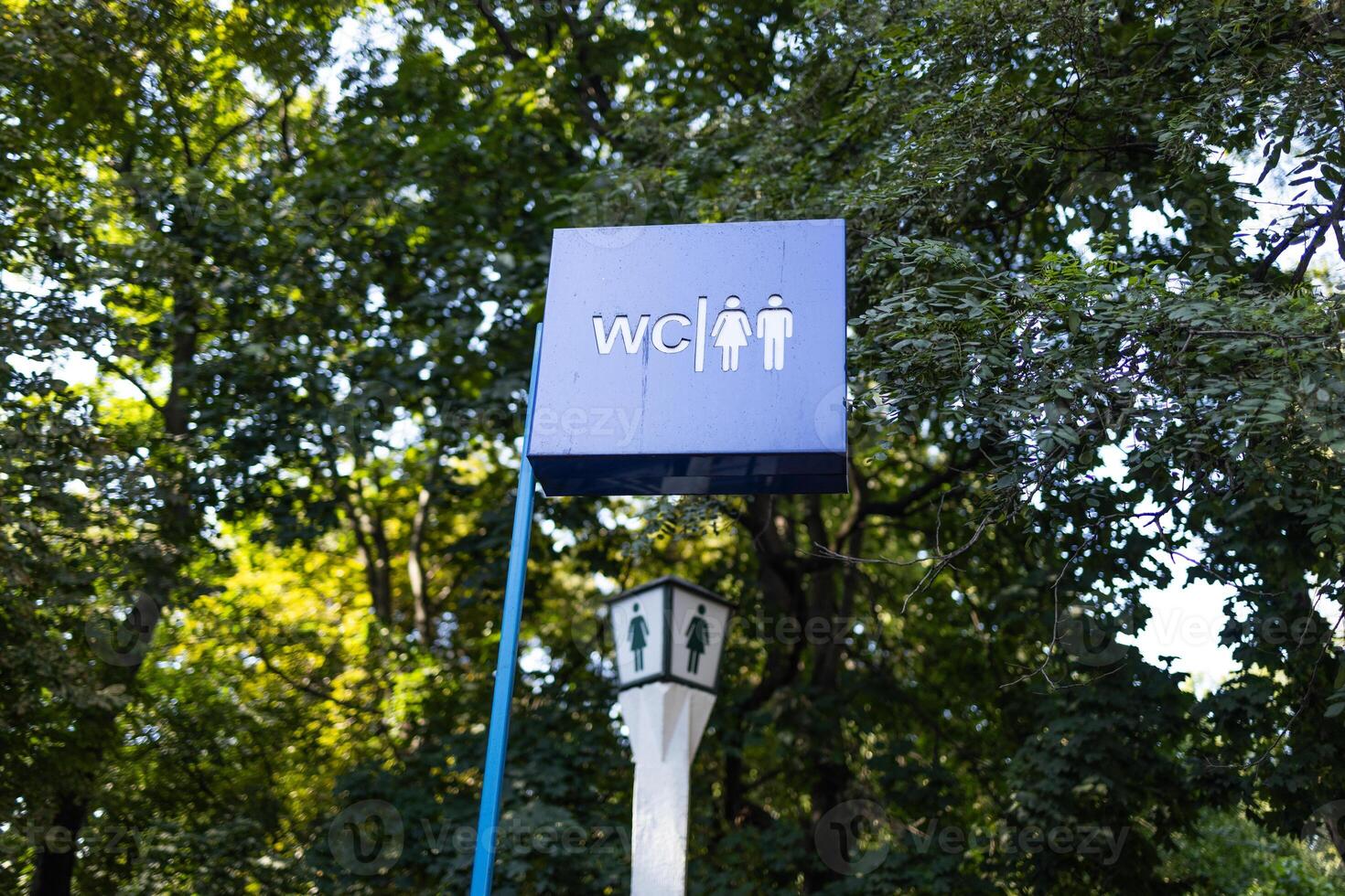 Toilet sign against the background of green trees in the park. White wc sign on blue metal plate photo