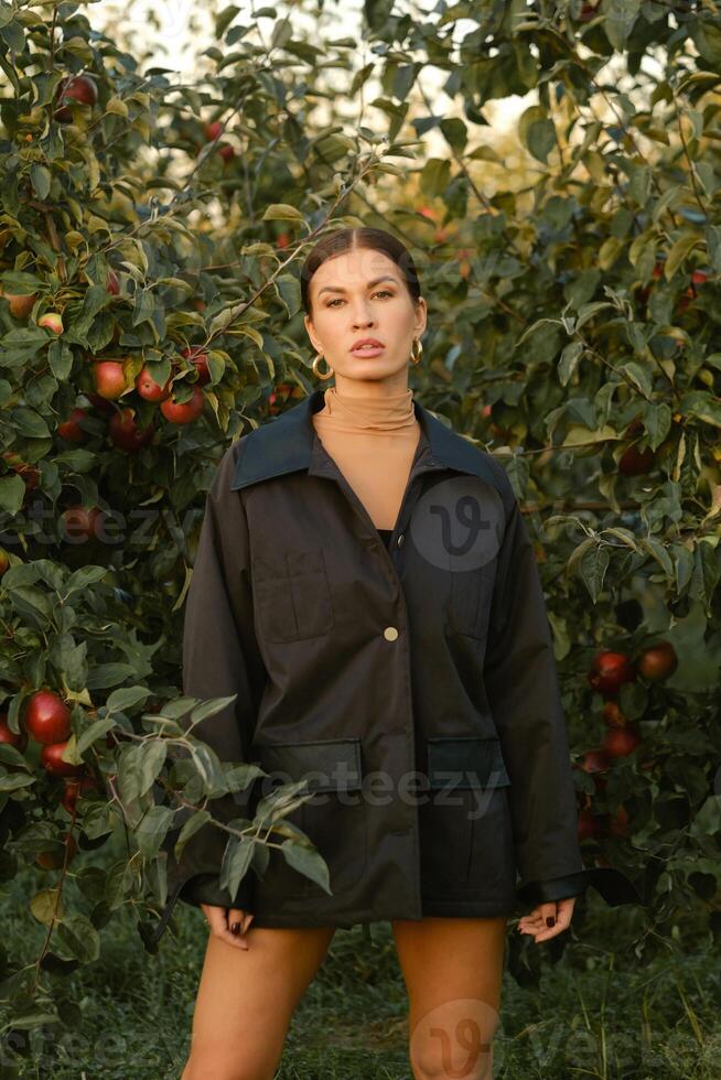 A beautiful girl in a black autumn jacket in an apple orchard. A fashionable girl at a fashion show in the apple orchard photo