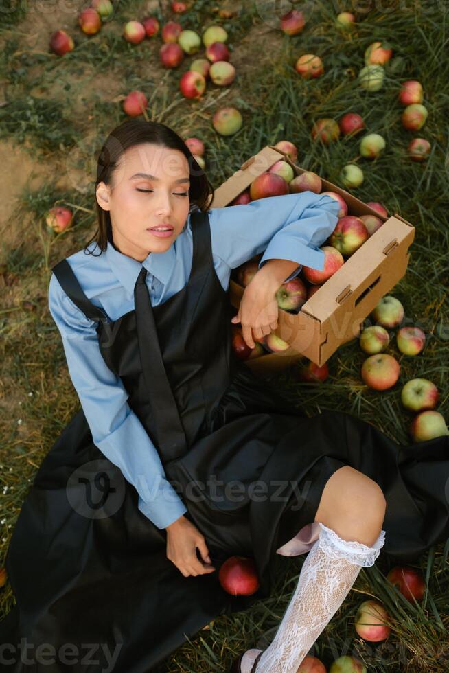 Portrait of a beautiful Asian woman in stylish clothes with apples in the garden. The girl is sitting on the ground and there are a lot of red apples around photo