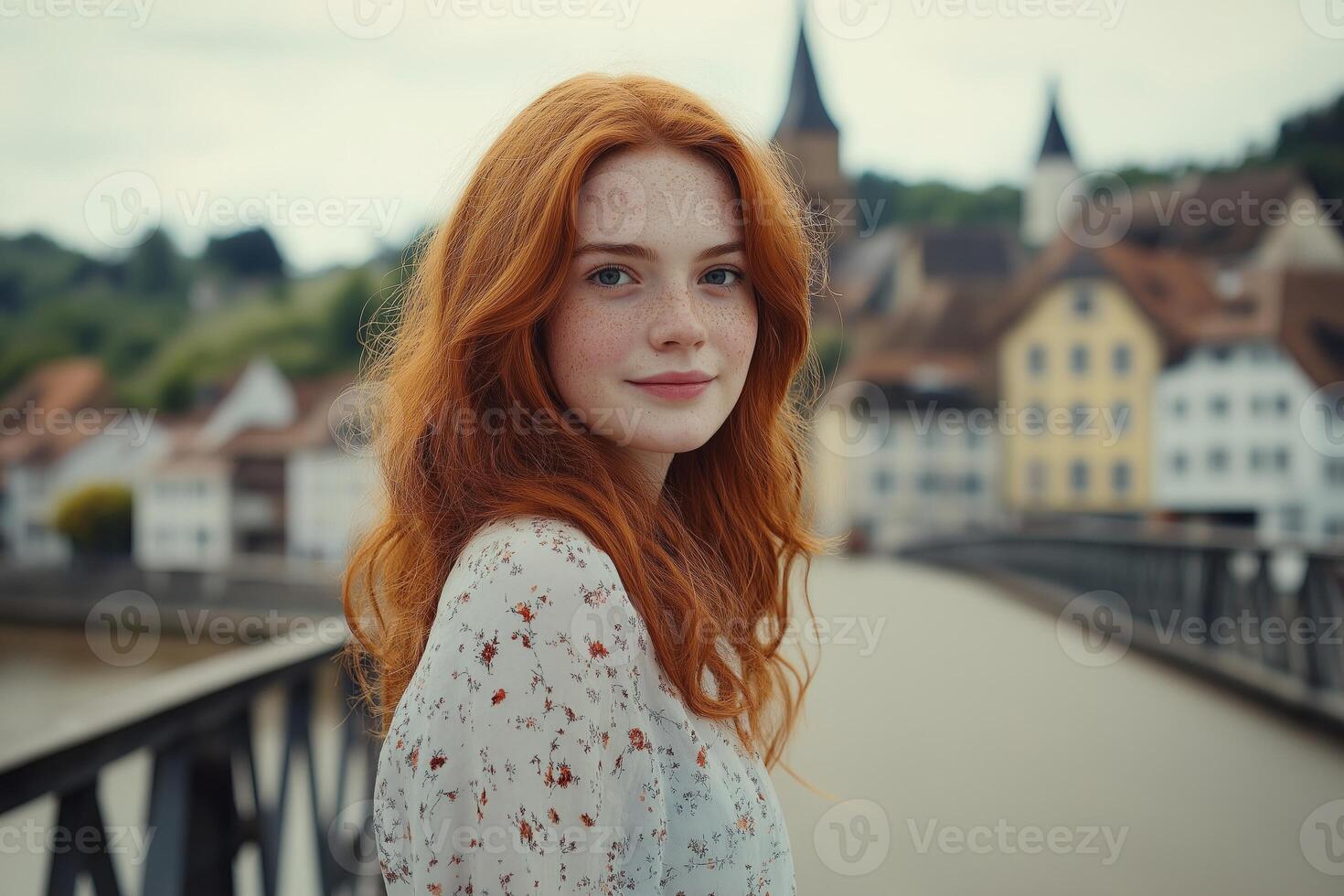 Young woman with red hair in a summer dress stands on a bridge overlooking a charming small town photo