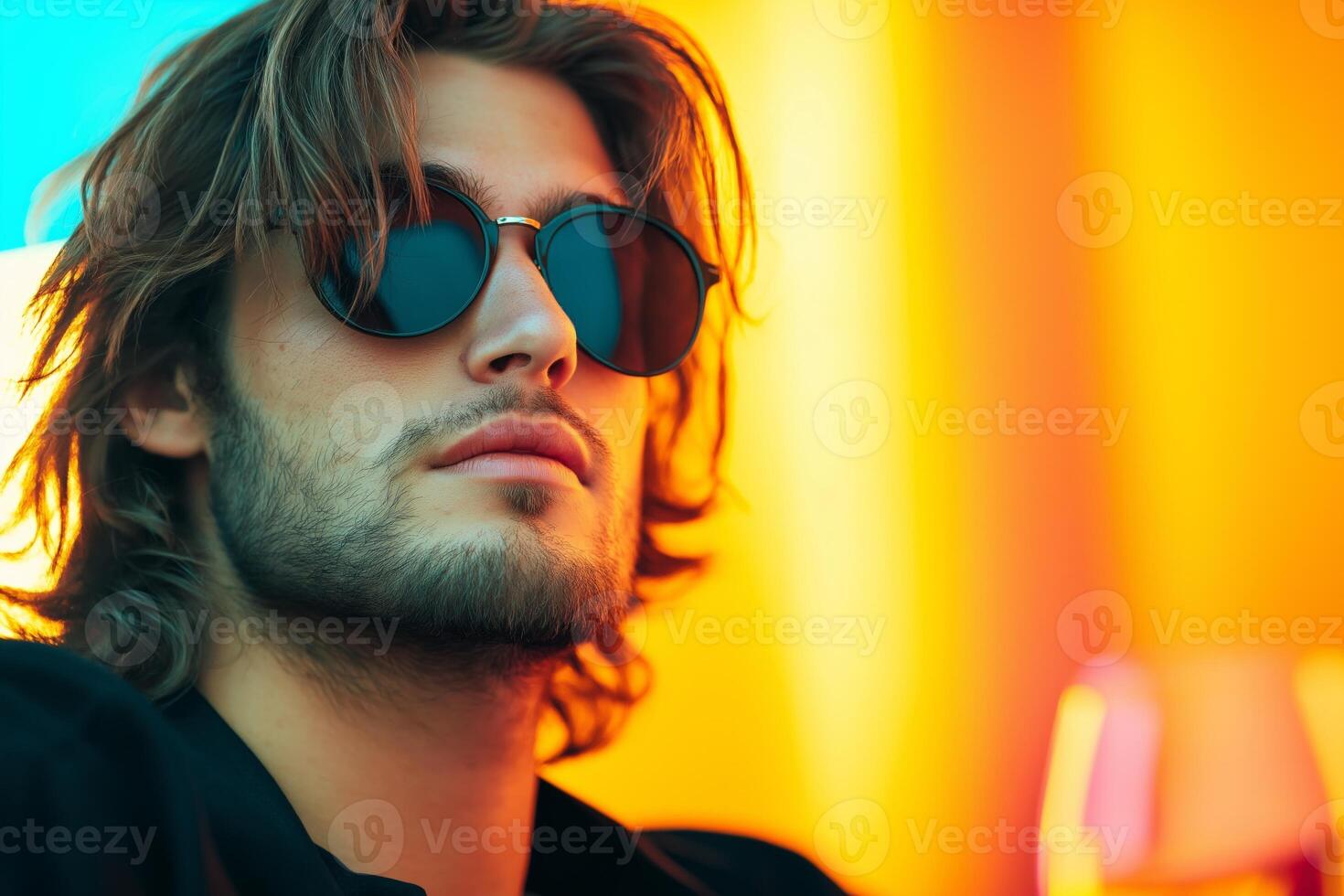 A stylish man with long hair and sunglasses looks contemplative in dark surroundings with vibrant colors photo
