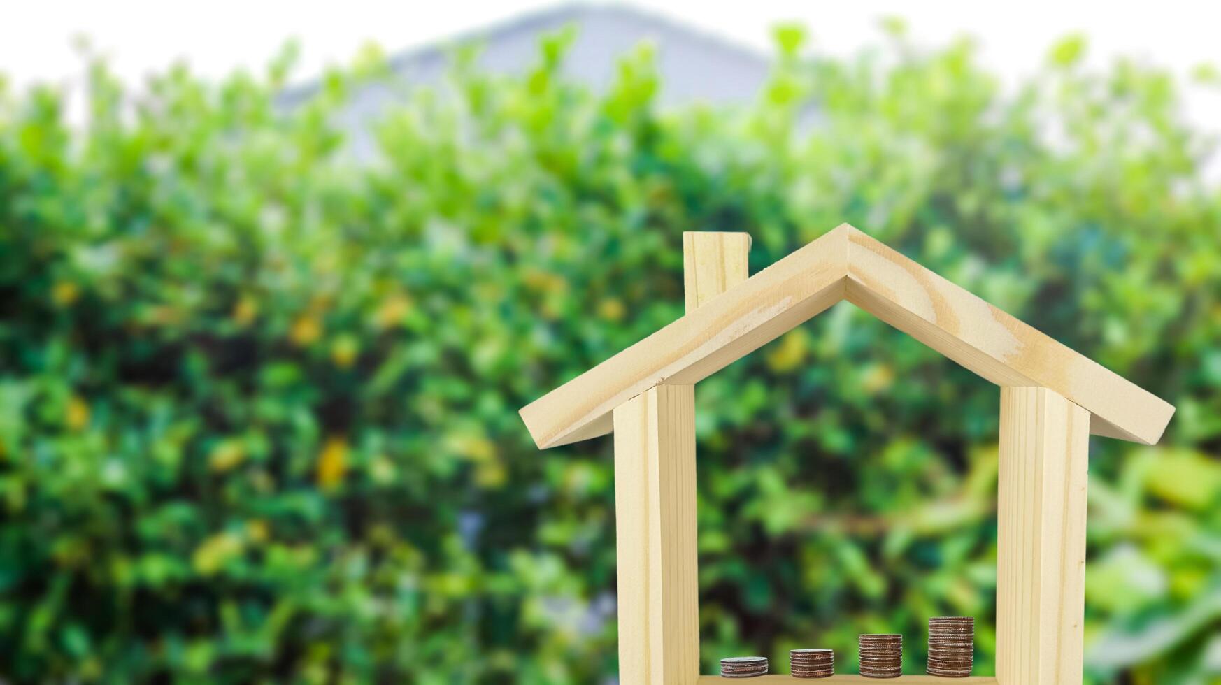 A wooden house model with coins arranged in ascending order inside, set against a blurred background of bushes, symbolizing financial growth and investment for home ownership. photo
