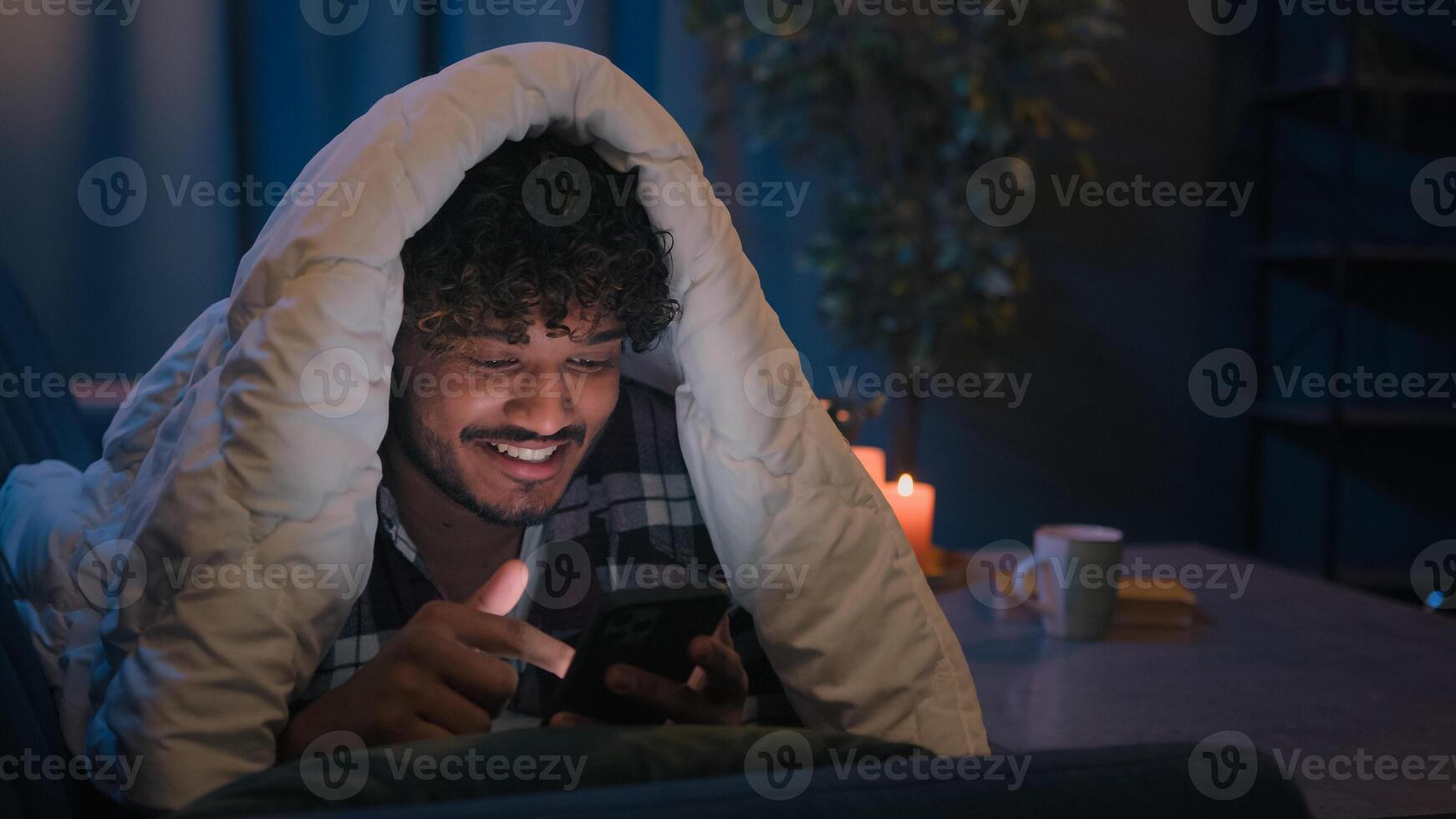 Smiling guy Indian man Arabian male at night evening home on couch under blanket cover with duvet looking mobile phone wow surprised amazed reading news gadget addict smartphone social media scrolling photo