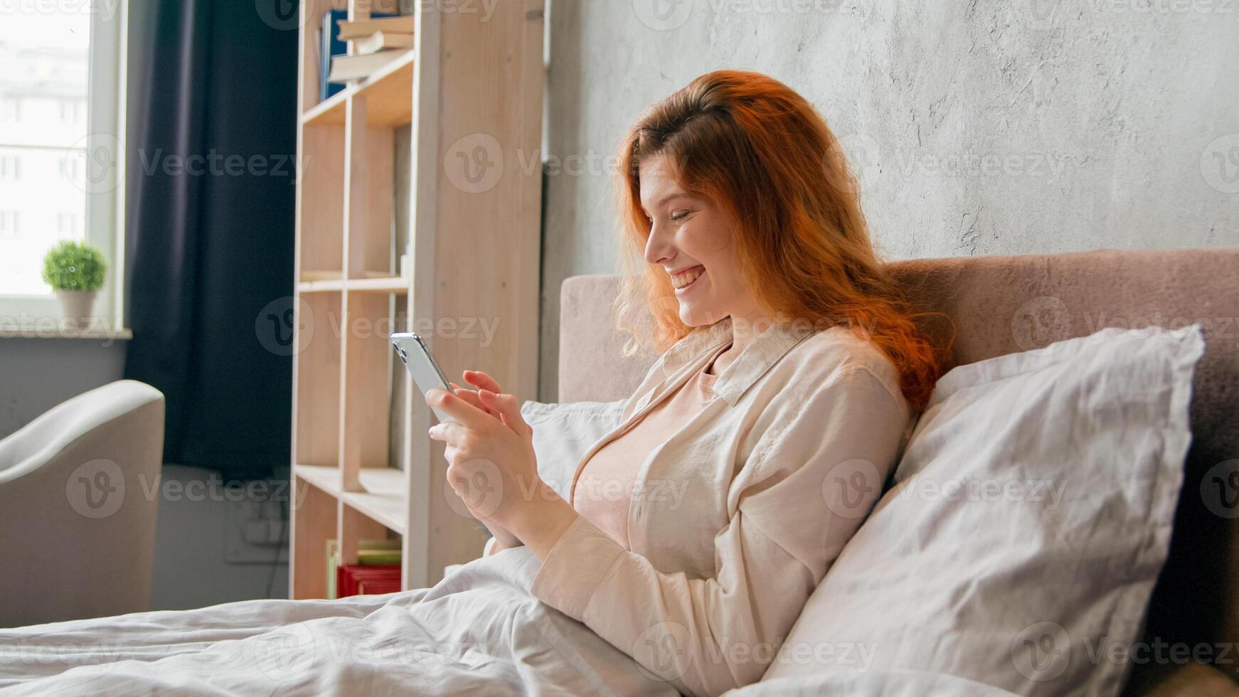 Relaxed Caucasian girl woman in bed at tranquil morning routine after wake up sleeping gadget internet addict female beauty influencer browsing mobile phone chatting in social media smiling laughing photo
