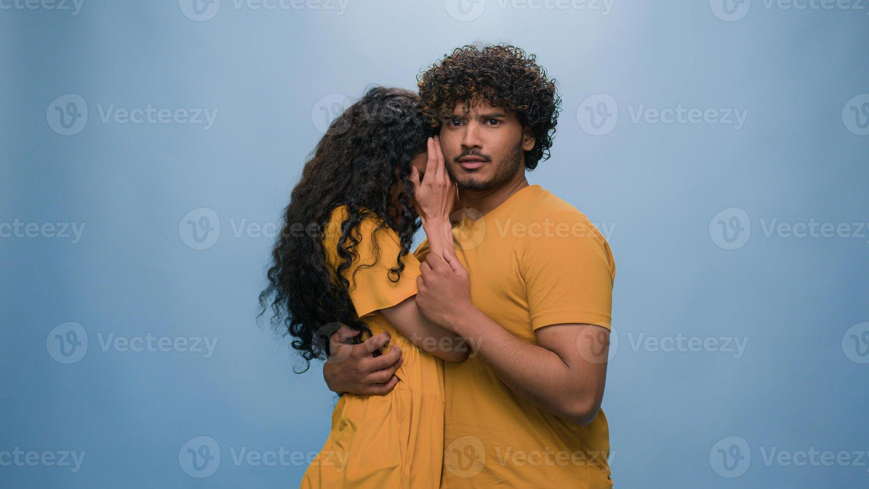 Multiracial shocked scared afraid couple amazed shocked Hispanic woman hiding behind Indian man astonishment wonder scare terrible horror ugly reaction blue studio background shock astonish fright photo
