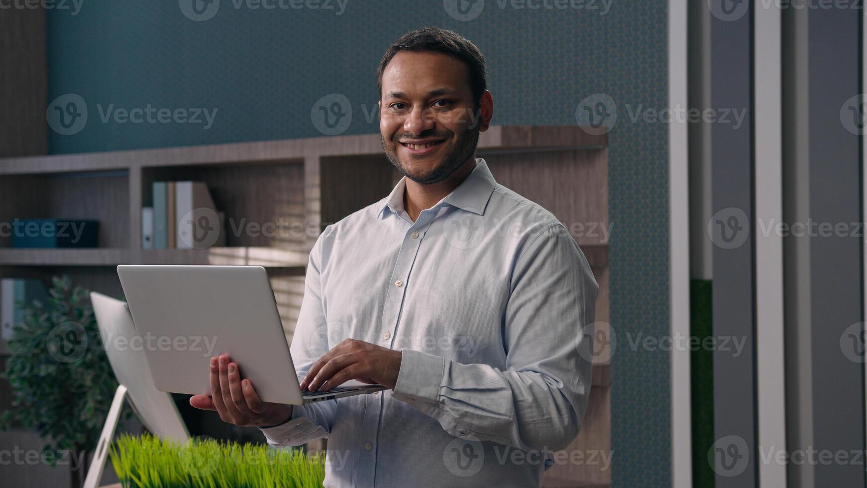 American male office worker standing at workplace in corporate company businessman working with computer man work online typing chatting with clients business task holding laptop smiling at camera photo