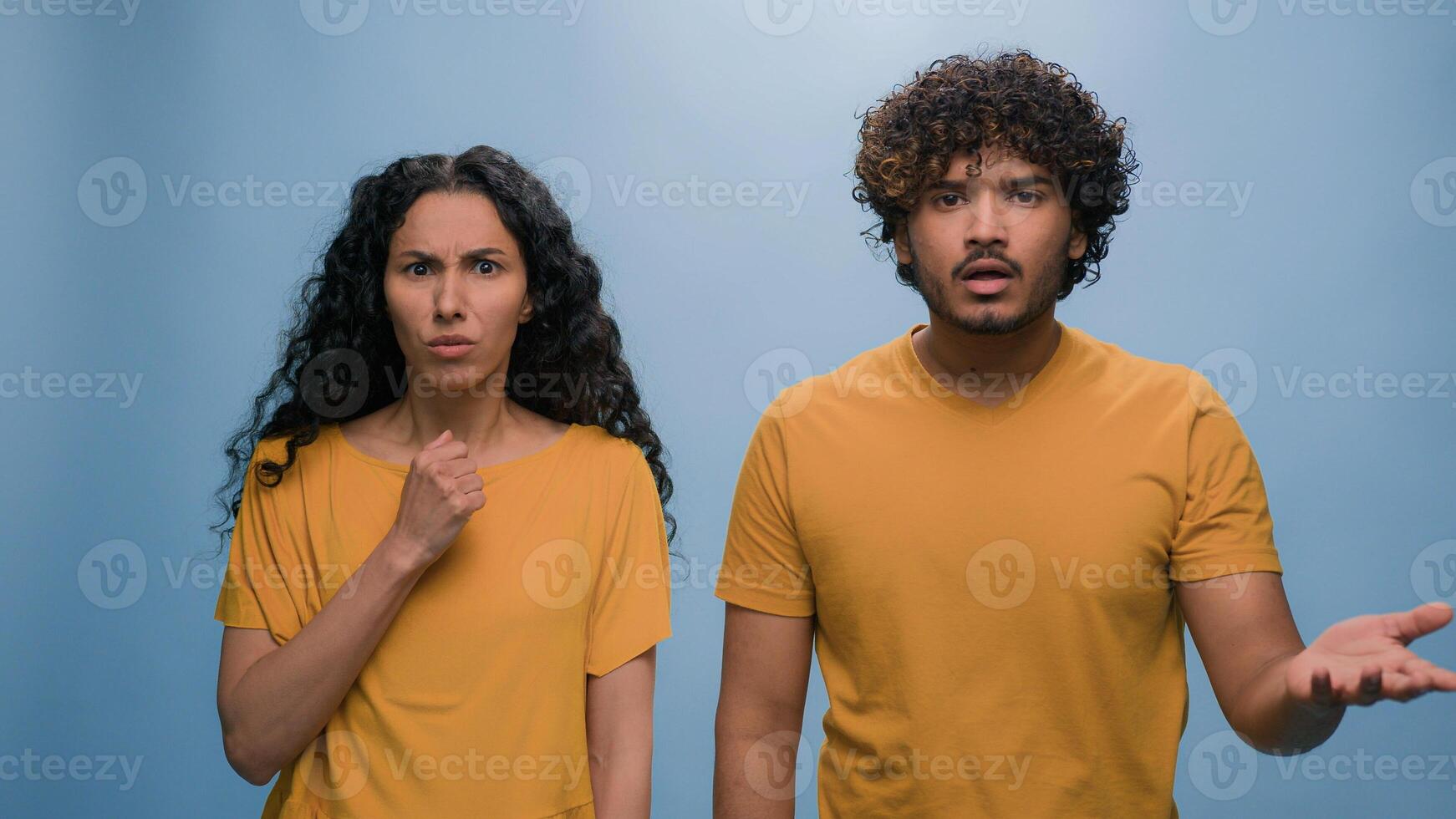 Multiracial shocked scared afraid couple amazed shocked Hispanic woman hiding behind Indian man astonishment wonder scare terrible horror ugly reaction blue studio background shock astonish fright photo