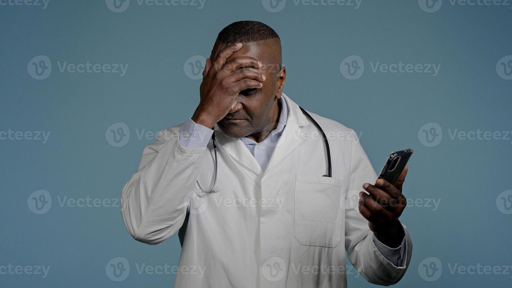Upset frustrated african american man doctor reading bad medical news on mobile phone in blue background receive notification worried male healthcare worker stressed over broken smartphone low battery photo
