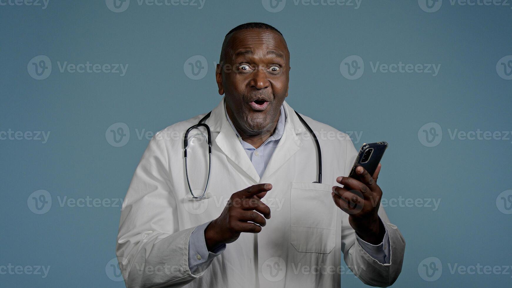 Happy African American man doctor male healthcare employee looking mobile phone screen receiving email reading good news excellent medical analysis result using smartphone in studio blue background photo