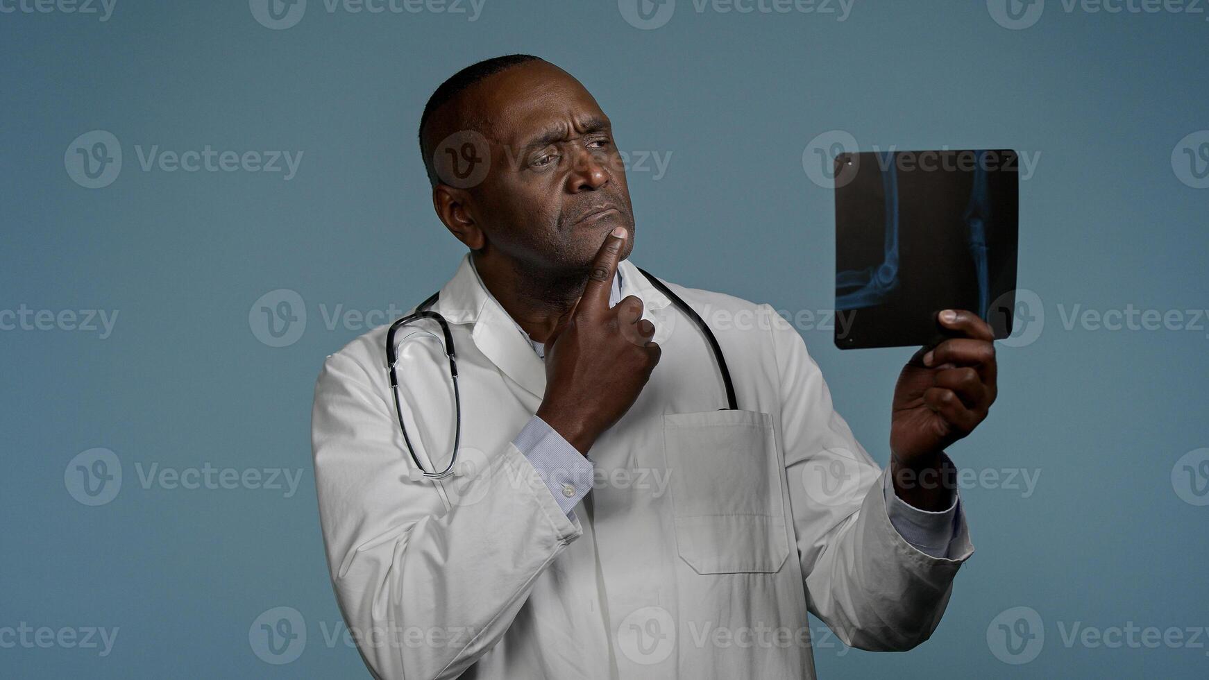 Pensive thoughtful african american man doctor ethnic male radiologist surgeon in studio blue background look at x-ray result checking radiography scan broken bone professional medic analyze trauma photo