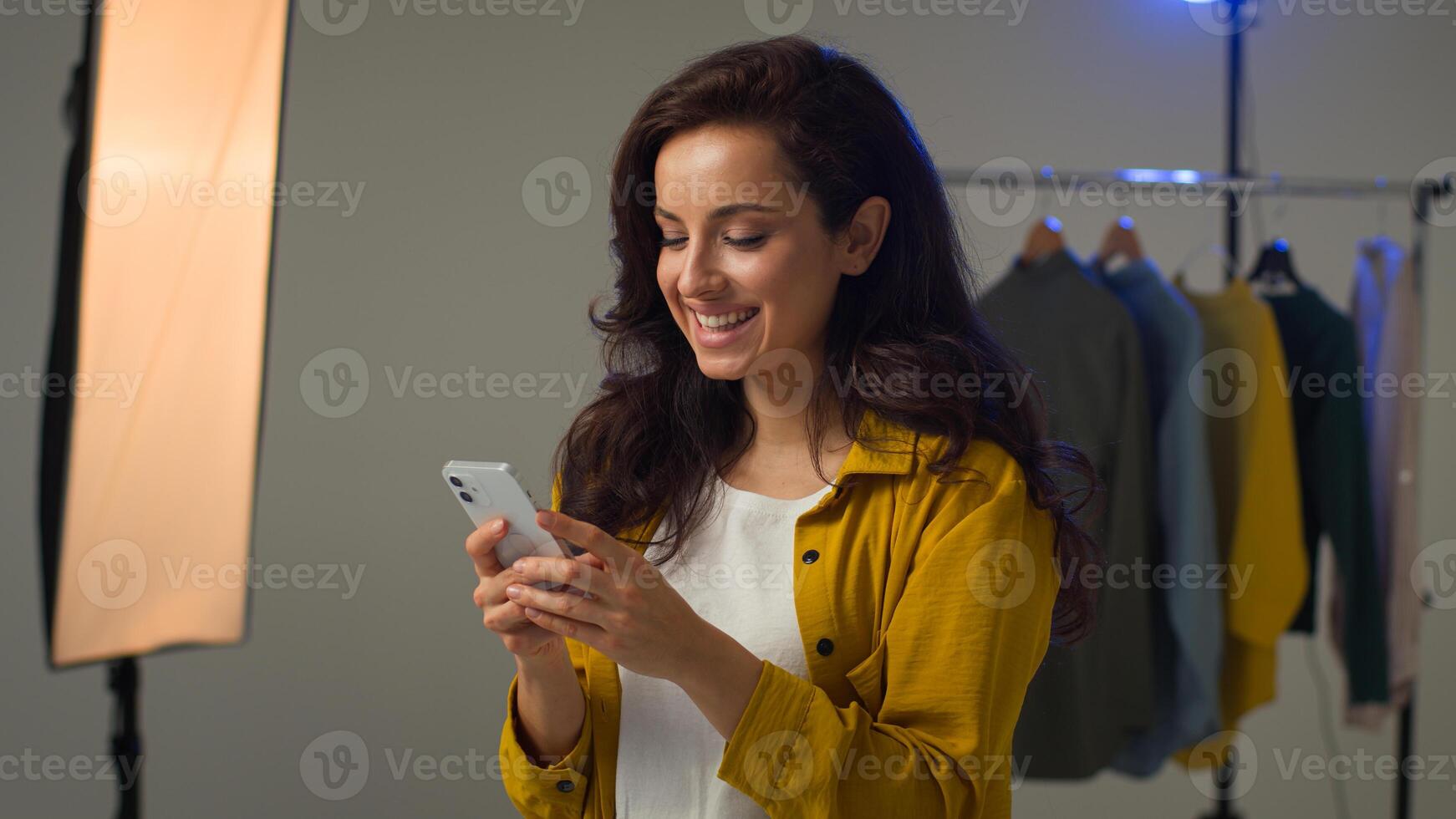 Smiling happy Caucasian woman model female blogger hold mobile phone smartphone looking screen scroll feed. Positive lady girl influencer chatting on social media blog browsing Internet web in studio photo