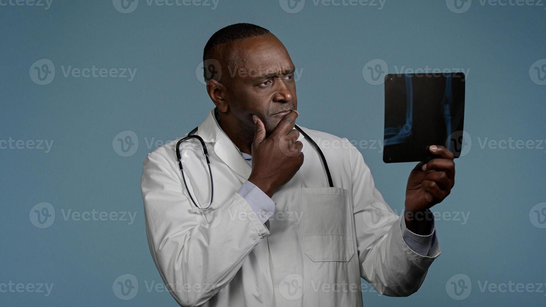 Pensive thoughtful african american man doctor ethnic male radiologist surgeon in studio blue background look at x-ray result checking radiography scan broken bone professional medic analyze trauma photo