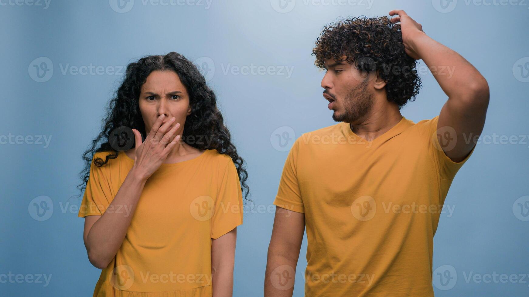 Multiracial shocked scared afraid couple amazed shocked Hispanic woman hiding behind Indian man astonishment wonder scare terrible horror ugly reaction blue studio background shock astonish fright photo