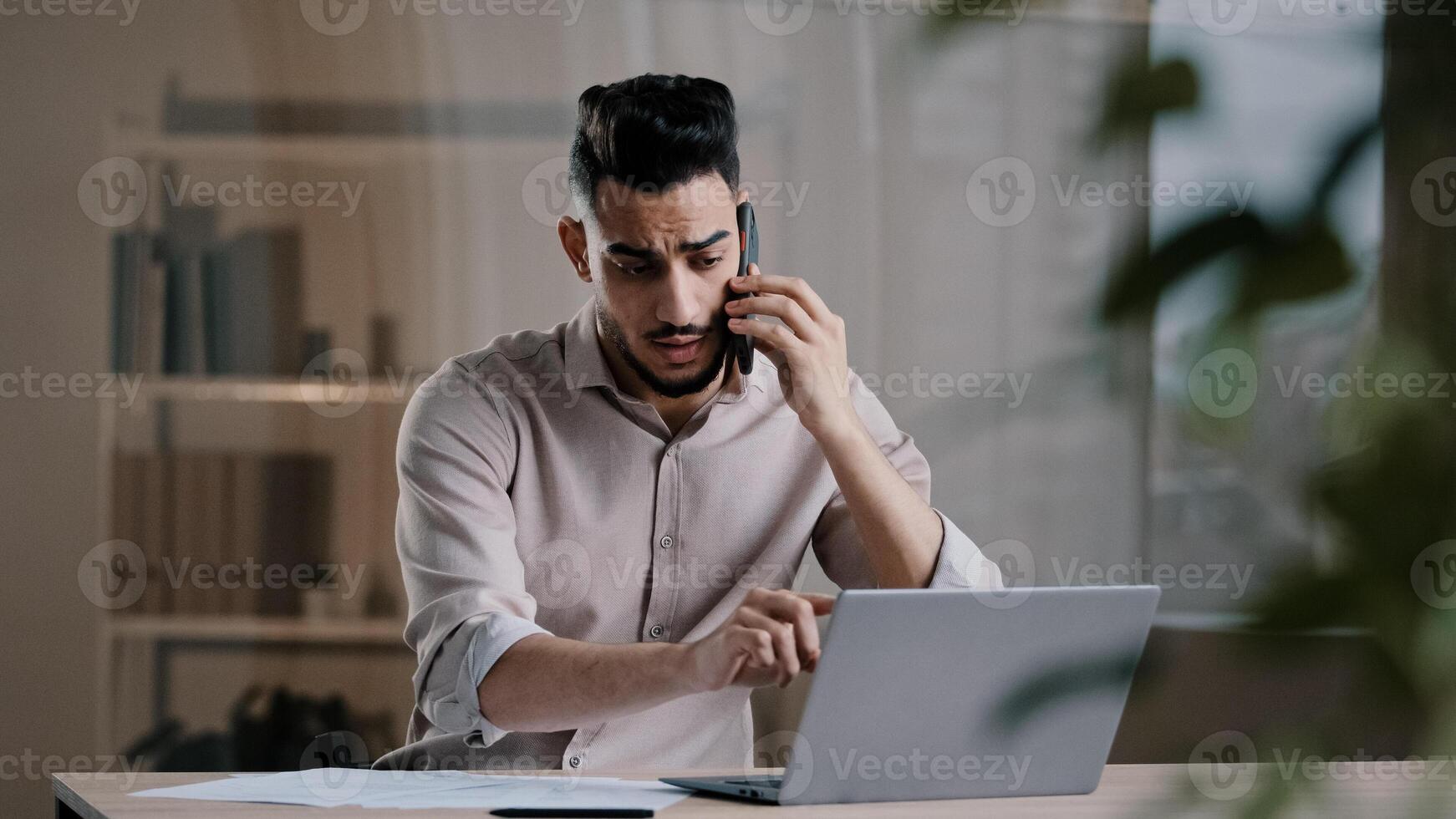 Busy millennial arabian business man web designer employee using laptop talking on cell phone involved in multitasking using gadgets discussing online project distant noting paper documents at office photo