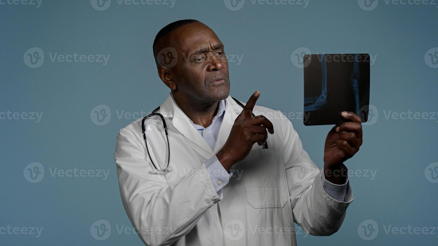 Pensive thoughtful african american man doctor ethnic male radiologist surgeon in studio blue background look at x-ray result checking radiography scan broken bone professional medic analyze trauma photo