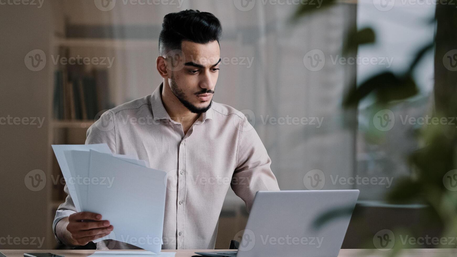 Serious arabian young business guy male professional worker employee type on computer keyboard copy paper text at electronic form focused hispanic man compare documents check online financial report photo