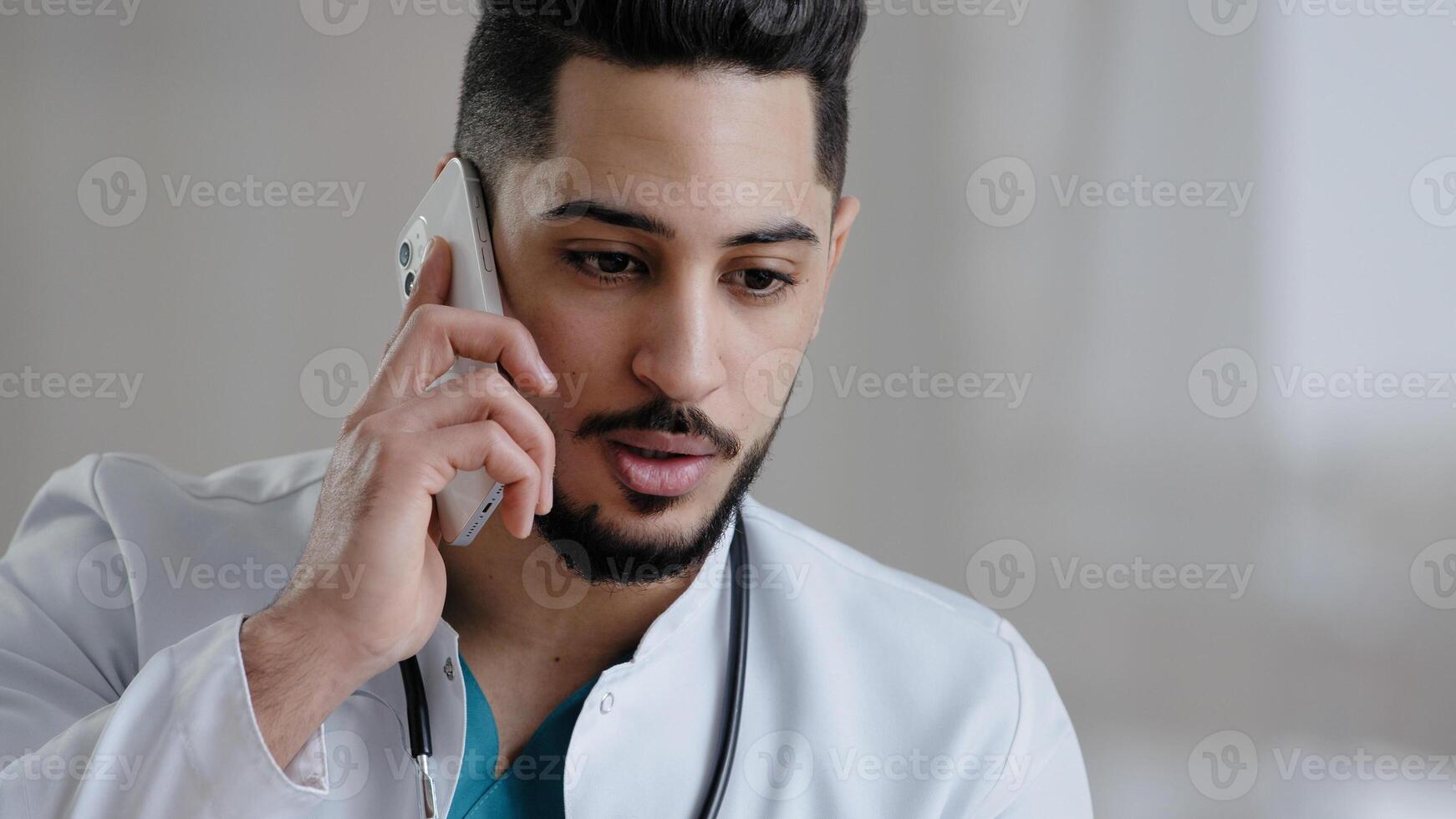 Serious male hispanic guy doctor therapist medical assistant consult patient distant discuss issues by mobile phone arab man adviser surgeon provide remote consultation use cellphone at clinic cabinet photo