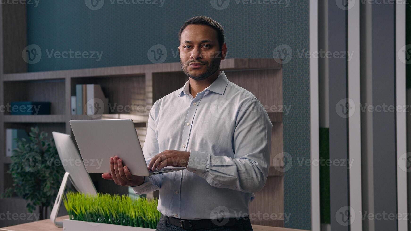 American male office worker standing at workplace in corporate company businessman working with computer man work online typing chatting with clients business task holding laptop smiling at camera photo