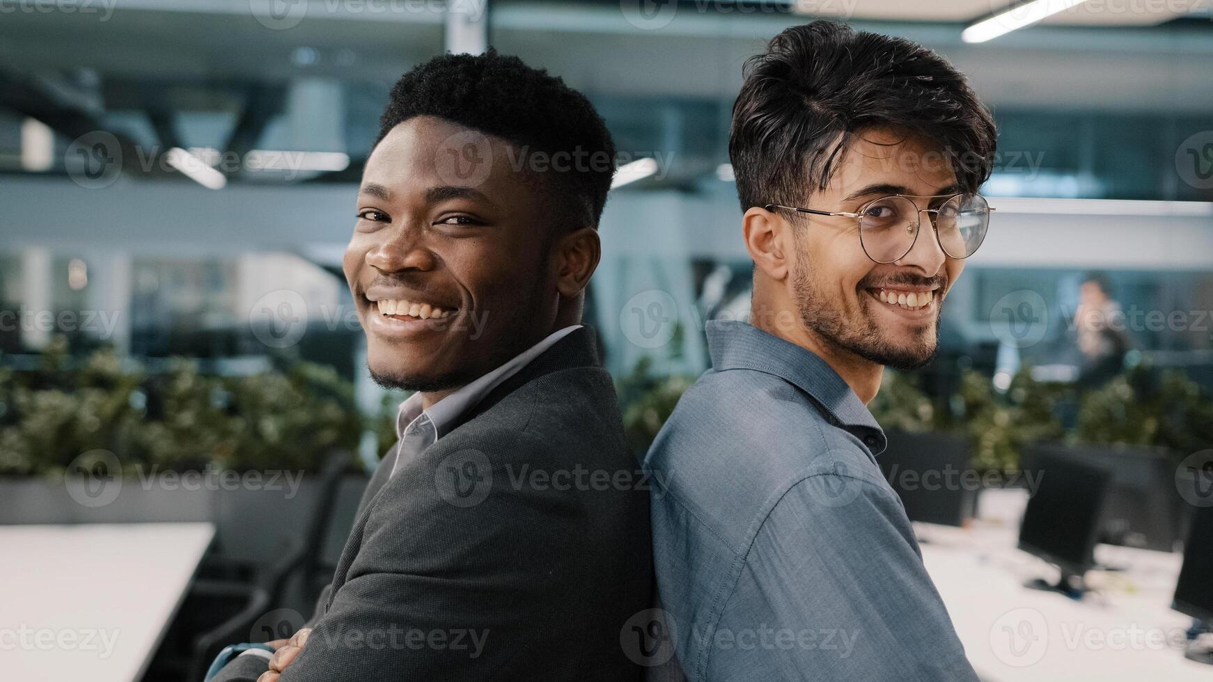 Portrait two multinational male colleagues men African man arab Indian manager posing in office company standing back to each other partnership looking at camera smiling business partners employees photo
