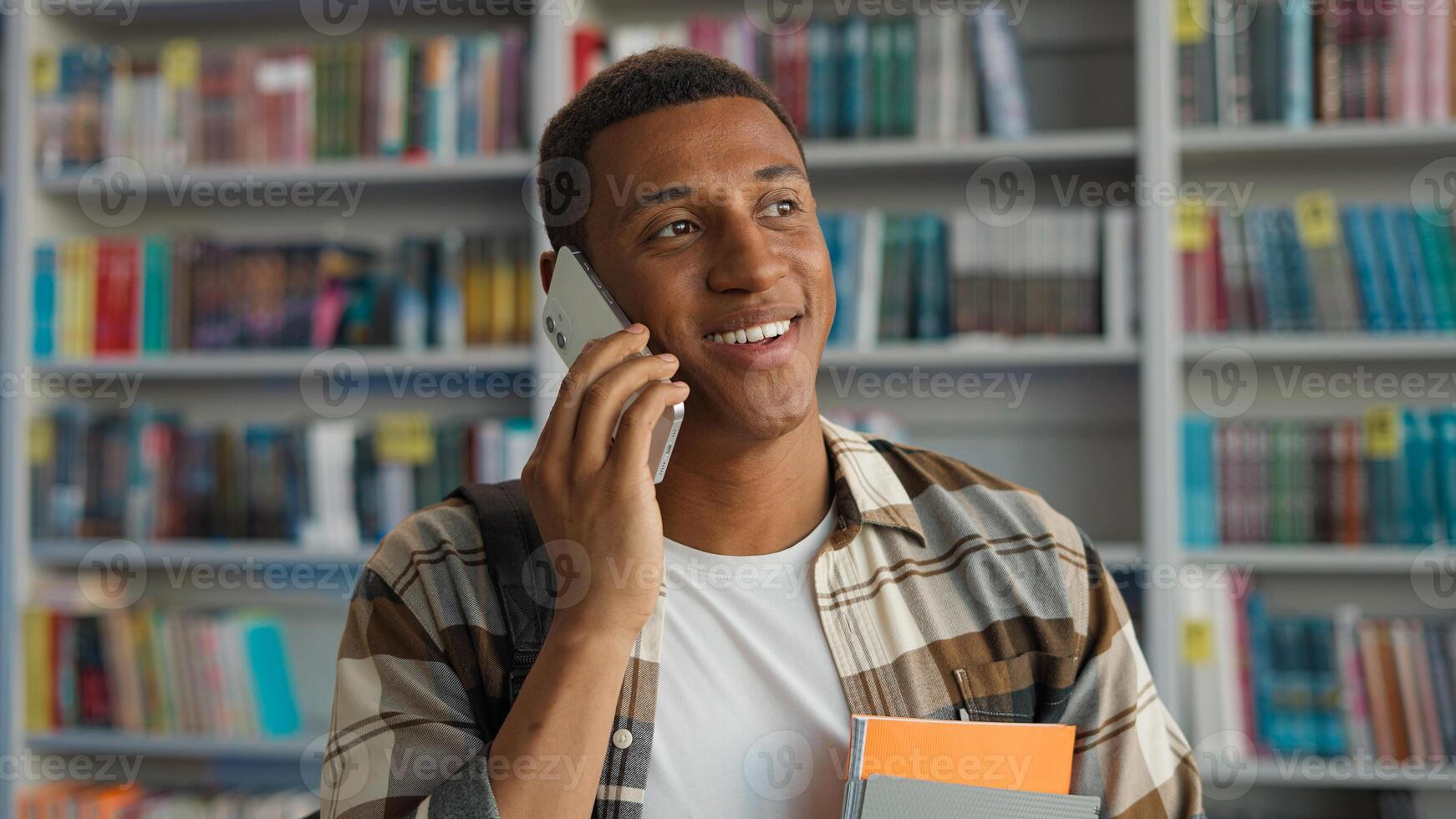 Happy African American man talking mobile phone smartphone technology communicate conversation library student learner university chatting smiling male guy bookshelf educational study calling laughter photo