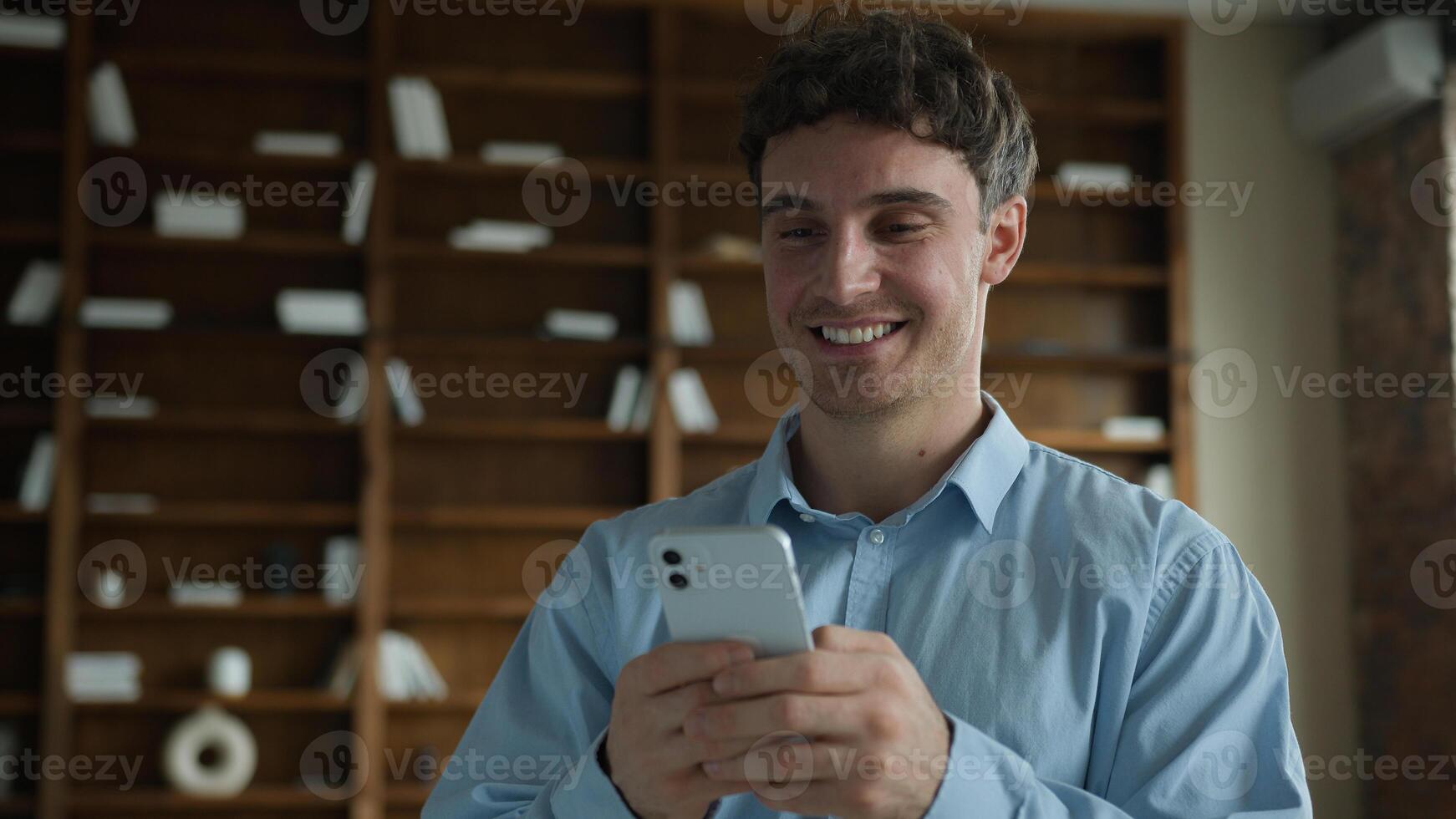 Happy wide smiling young Caucasian male business man businessman guy entrepreneur using mobile phone smartphone typing message chatting browsing internet enjoy online app game ordering delivery indoor photo