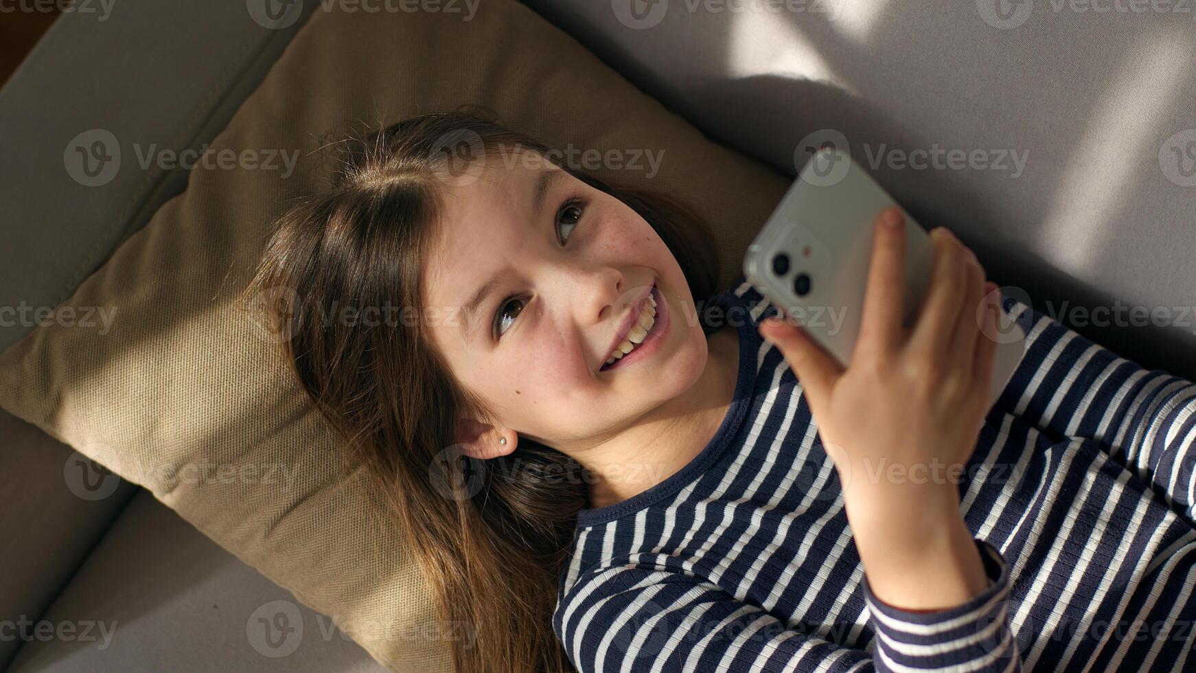 Caucasian European little girl lying on sofa holding using mobile phone smartphone child gadget online texting sms social networks smiling laughing enjoyment watching funny internet chatting kid photo